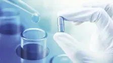 Gloved hand holding a capsule near a test tube as a drop from a pipette falls, illustrating pharmaceutical research.